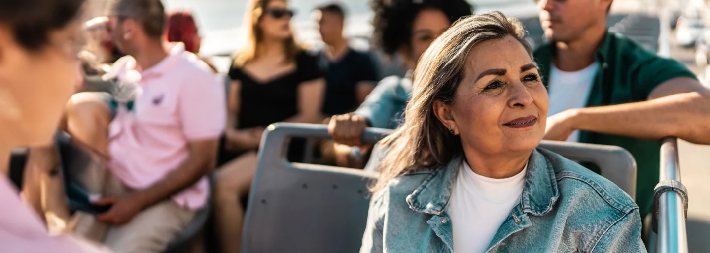 A woman with long gray hair sits on an open-air bus, gazing into the distance.